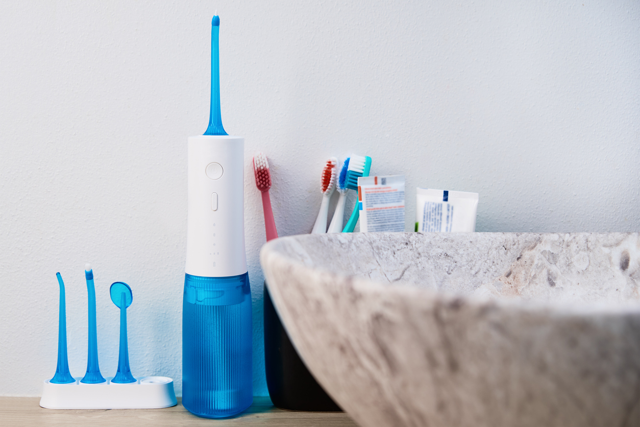 The image shows a Waterpik beside a family set of toothbrushes and a bathroom sink. The image represents the question of whether Waterpik is good for cleaning veneers.